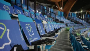 Plan rapproché sur un siège des gradins vides de l'Arena Stade couvert de Liévin. Des drapeaux et tote-bags sont déposés sur chacun des sièges bleus de la tribune.