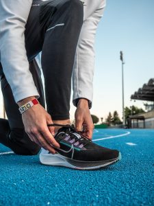 Zoom sur les pieds d'un homme faisant ses lacets, avec une paire de Nike Zoom Pegasus 38 au pied. Il est sur une piste d'athlétisme en tartan bleu. Il y a une tribune de stade en arrière plan.